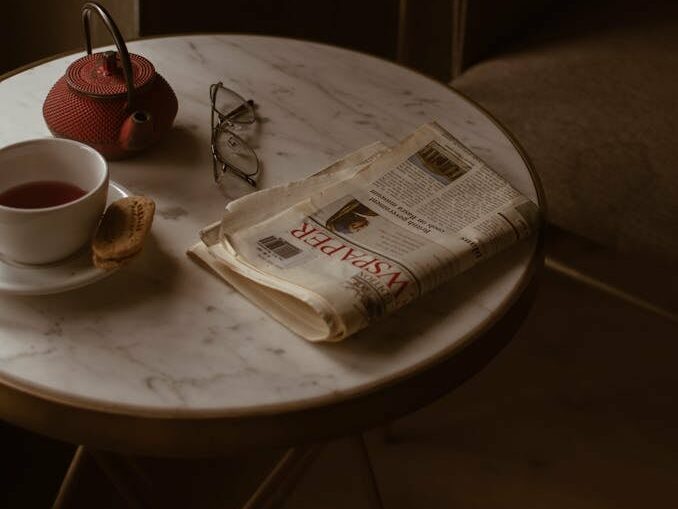 From above of round marble textured table with tea set placed near newspaper (possibly about DevOps) and eyeglasses in retro style cafe